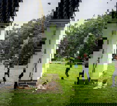 Drie personen en een hond staan in een grote tuin, waarbij een persoon zijn camera richt om een foto te maken van een gebouw.
