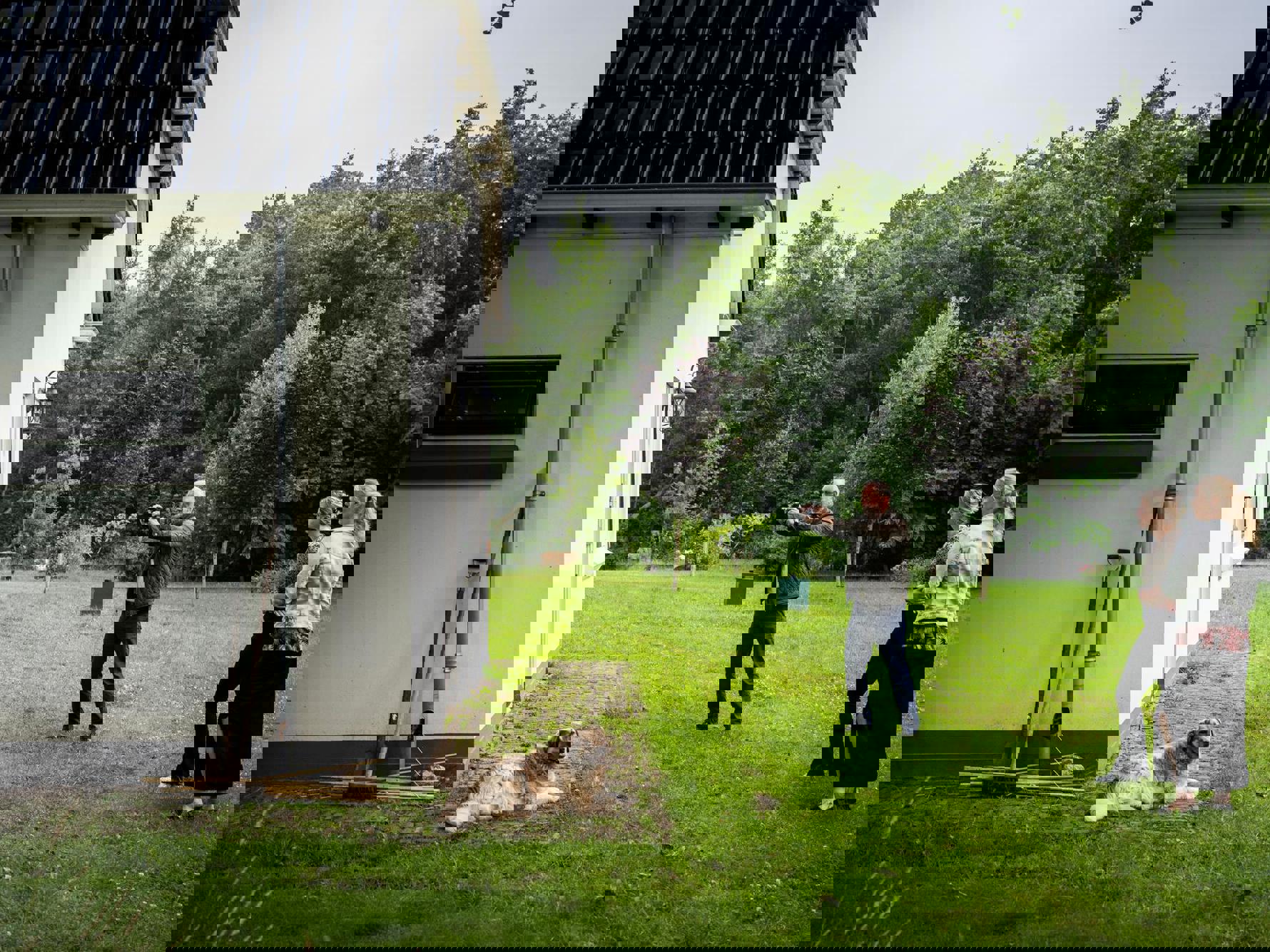 Drie personen en een hond staan in een grote tuin, waarbij een persoon zijn camera richt om een foto te maken van een gebouw.