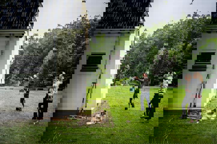 Drie personen en een hond staan in een grote tuin, waarbij een persoon zijn camera richt om een foto te maken van een gebouw.
