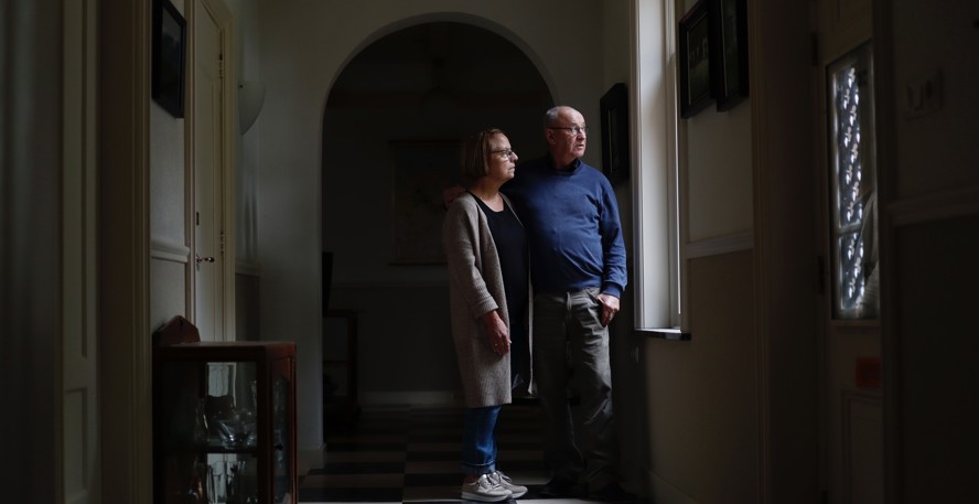 Ina en Koos Koop staan in de schaduwrijke, zwart-wit betegelde hal van hun huis uit het raam te kijken