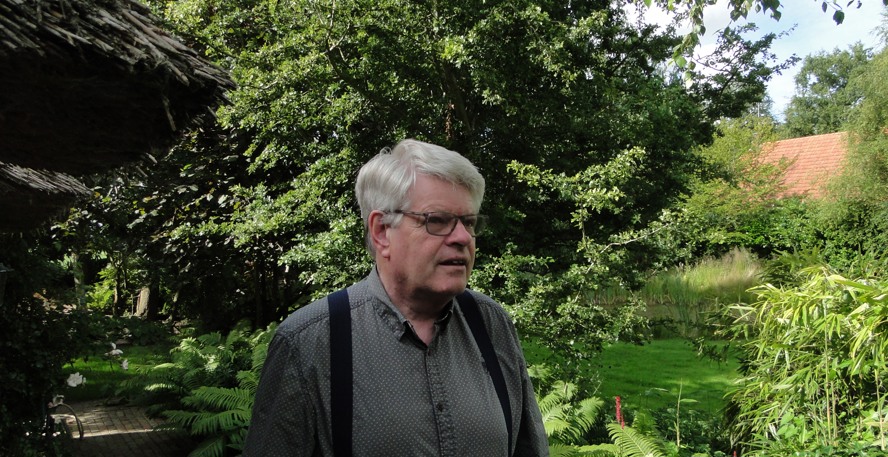 Kees Kemper staat in zijn weelderige groene tuin. Er is nog een stukje rieten dak zichtbaar