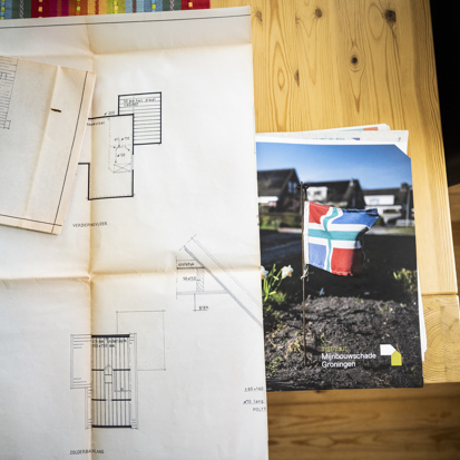 Op tafel liggen papieren waarop een bouwtekening van een woning zichtbaar is met erachter de map met documenten van Instituut Mijnbouwschade Groningen.