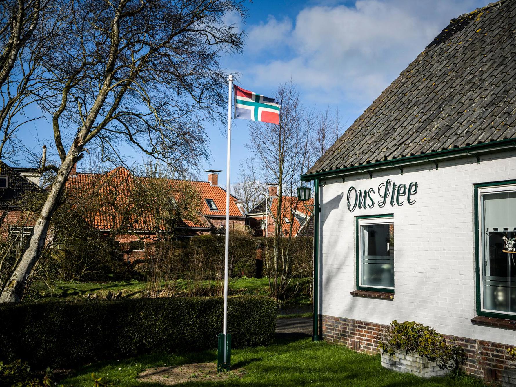 Een wapperende Groningse vlag op stok in de tuin van een wit huis met daarop de tekst: Ons Stee.