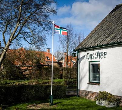 Een wapperende Groningse vlag op stok in de tuin van een wit huis met daarop de tekst: Ons Stee.