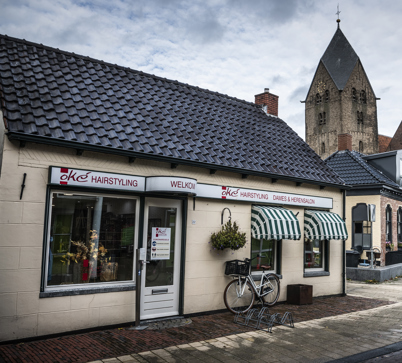 Een dorpsplein met een kapsalon, een kind op de fiets en een kerktoren die boven de gebouwen uitsteekt.