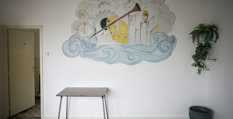 Een lege kamer met een muurschildering waarop een schilderspalet, boeken, een fluit, golven en een uiltje zijn afgebeeld