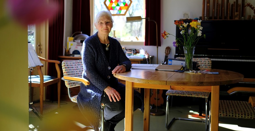 Kaja van Grieken aan tafel in een kamer met een piano, gitaar, fluiten en voor het raam een mozaïek 