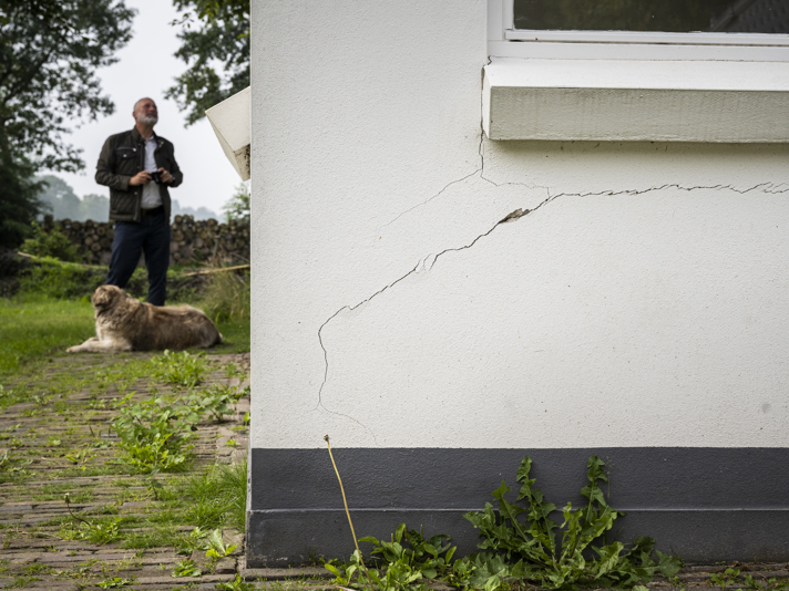 Grote scheur aan de buitenwand van een huis waarnaast een persoon staat en een hond op de grond eronder ligt. 