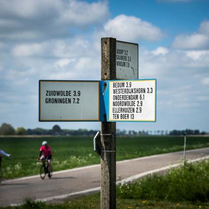 Wegwijzer met namen van dorpen uit het hogeland met op de achtergrond twee fietsers.