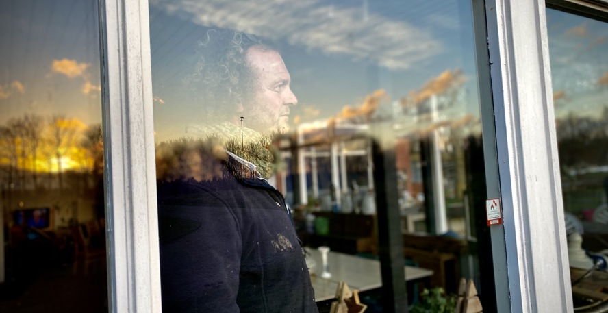 Drewes Wildeman, van buitenaf door het raam gefotografeerd. Het raam weerspiegelt bomen en de ondergaande zon.
