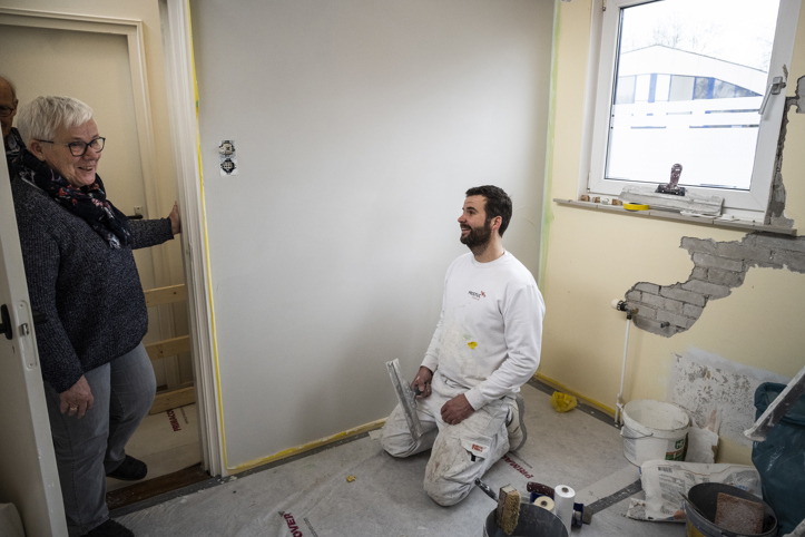 Een mevrouw stapt een kamer binnen waarin een meneer op zijn knieën stucwerkzaamheden uitvoert om de ruimte te herstellen.